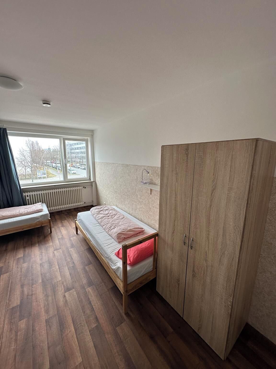 A small, well-lit room with a single bed, a wooden wardrobe, and a window with a view of the outside.