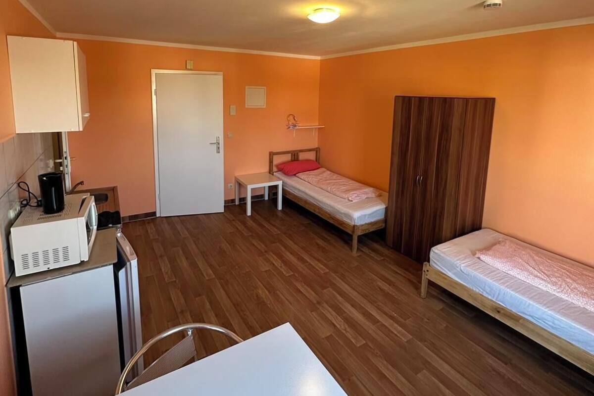 Una habitación compacta con dos camas individuales, un pequeño rincón americano, una mesa blanca y una puerta.
