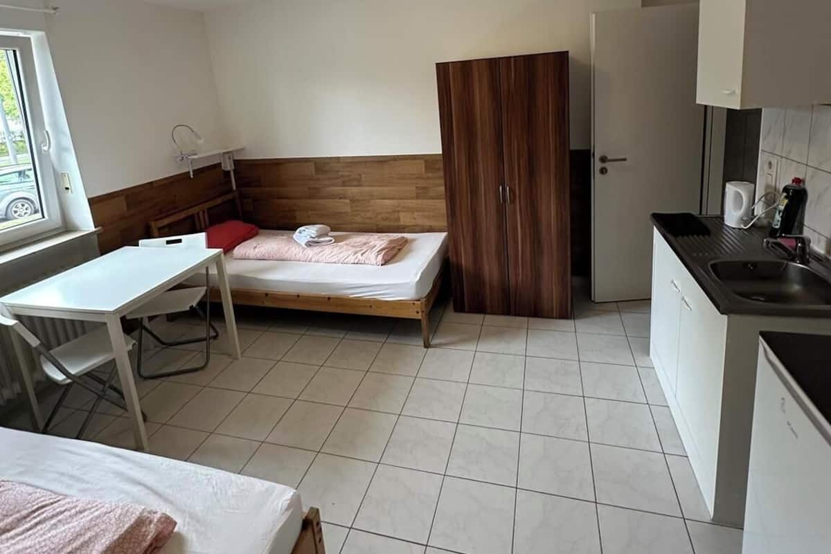Una habitación pequeña con cama, escritorio, kitchenette y armario.