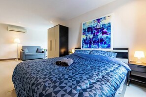 Une chambre d’hôtel moderne avec un grand lit, une couvre-lit bleue à motifs géométriques, une grande peinture abstraite au mur, un fauteuil bleu et une table de chevet avec une lampe.