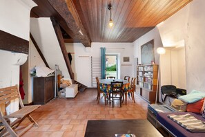 Een knus vertrek met houten plafond, een eettafel met stoelen, een bank, een boekenkast en een raam met gordijnen.