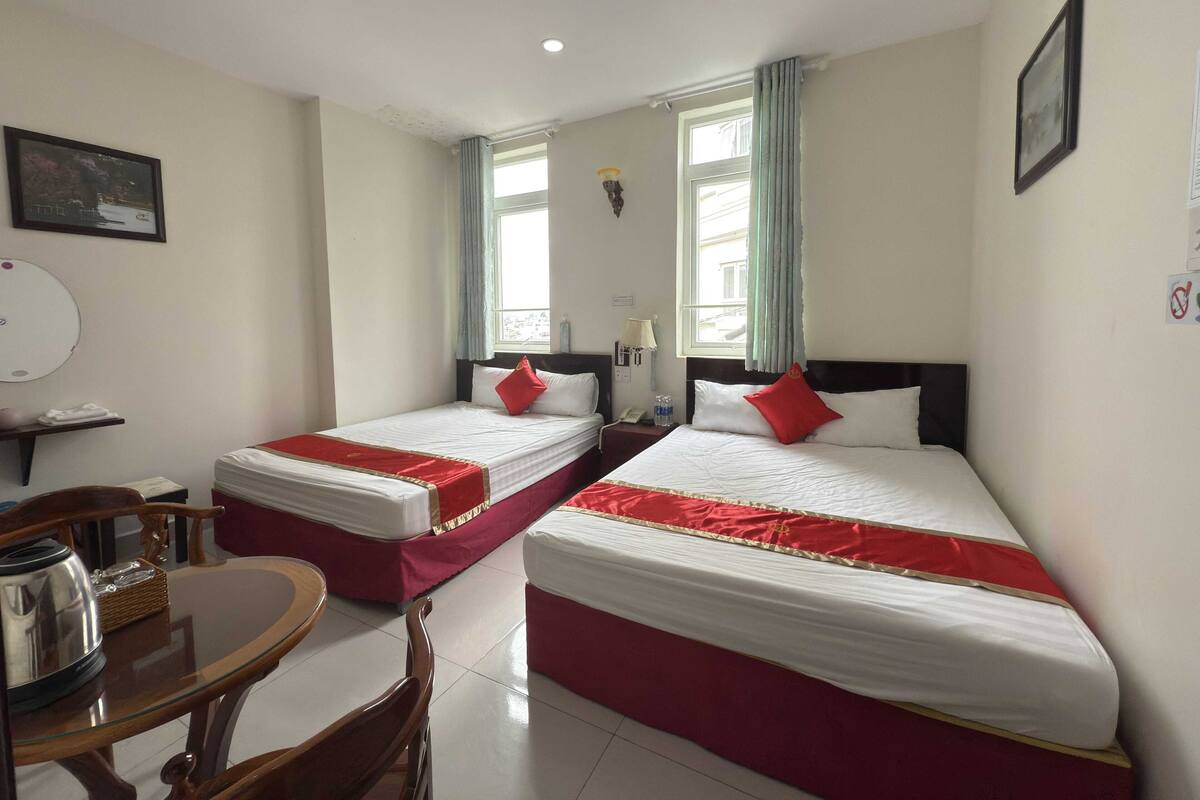 Ein Hotelzimmer mit zwei Betten, einem kleinen Tisch, Stühlen, einem Fenster mit Vorhängen und einer Wandlampe.