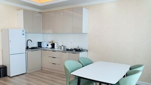 Une cuisine moderne avec un réfrigérateur blanc, un four à micro-ondes, un grille-pain et une cuisinière. Les armoires sont de couleur beige et le plan de travail est blanc. Une table entourée de quatre chaises vertes se trouve au premier plan.