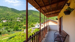 A balcony with a stone floor, a wooden bench, and a view of a green valley with houses.