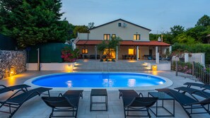 Una piscina con sillones de descanso, una casa con techo de tejas rojas y un muro de piedra.