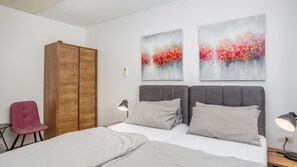 Un dormitorio con una cama, dos cabeceras grises, un armario de madera, una silla roja y dos cuadros colgados en la pared.