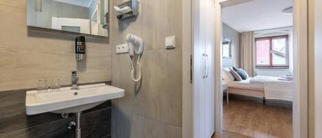 Un baño moderno con lavamanos, espejo, secador de pelo y toallero. Se ve un dormitorio al fondo con cama y ventana.