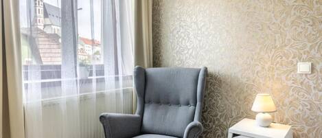 A room with a grey armchair, a white bedside table with a lamp, and a patterned wallpaper.