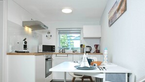Een compacte keuken met witte kastjes, een magnetron, een koffiezetapparaat en een raam met uitzicht op groen.