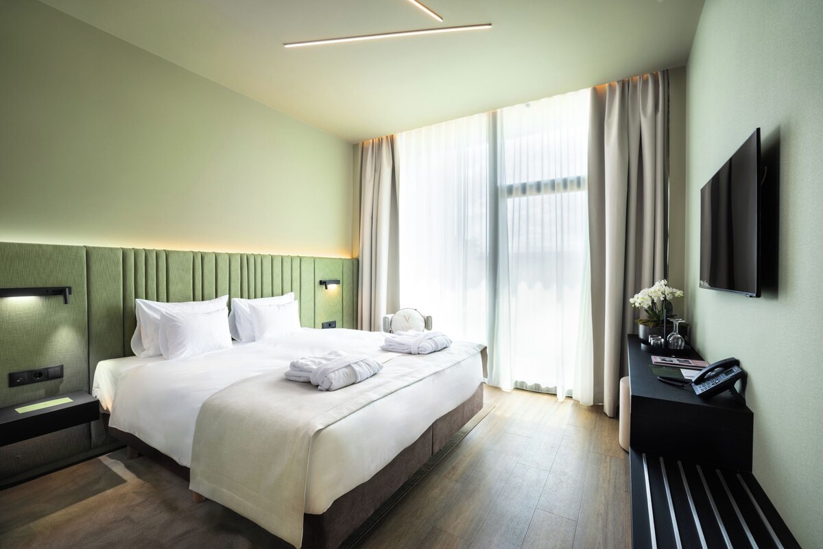 Ein Hotelzimmer mit einem großen Bett, Nachttischen, einem Schreibtisch mit Stuhl, einem Flachbildfernseher und einem großen Fenster mit Vorhängen.