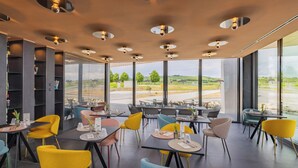 Ein modernes Restaurant mit großen Fenstern, das einen Blick auf eine Straße und Grünflächen bietet. Tische sind mit gelben und blauen Stühlen gedeckt.