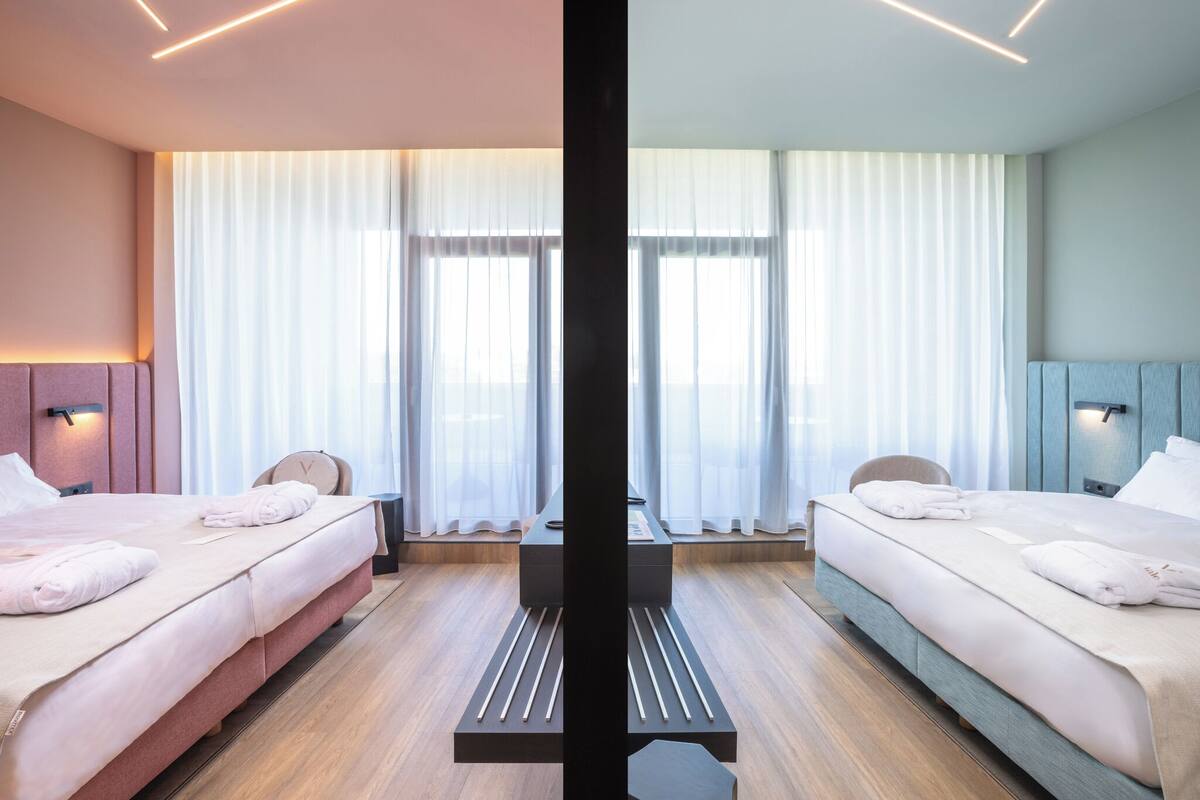 Habitación de hotel moderna con dos camas, una mesa central y un amplio ventanal con cortinas translúcidas.