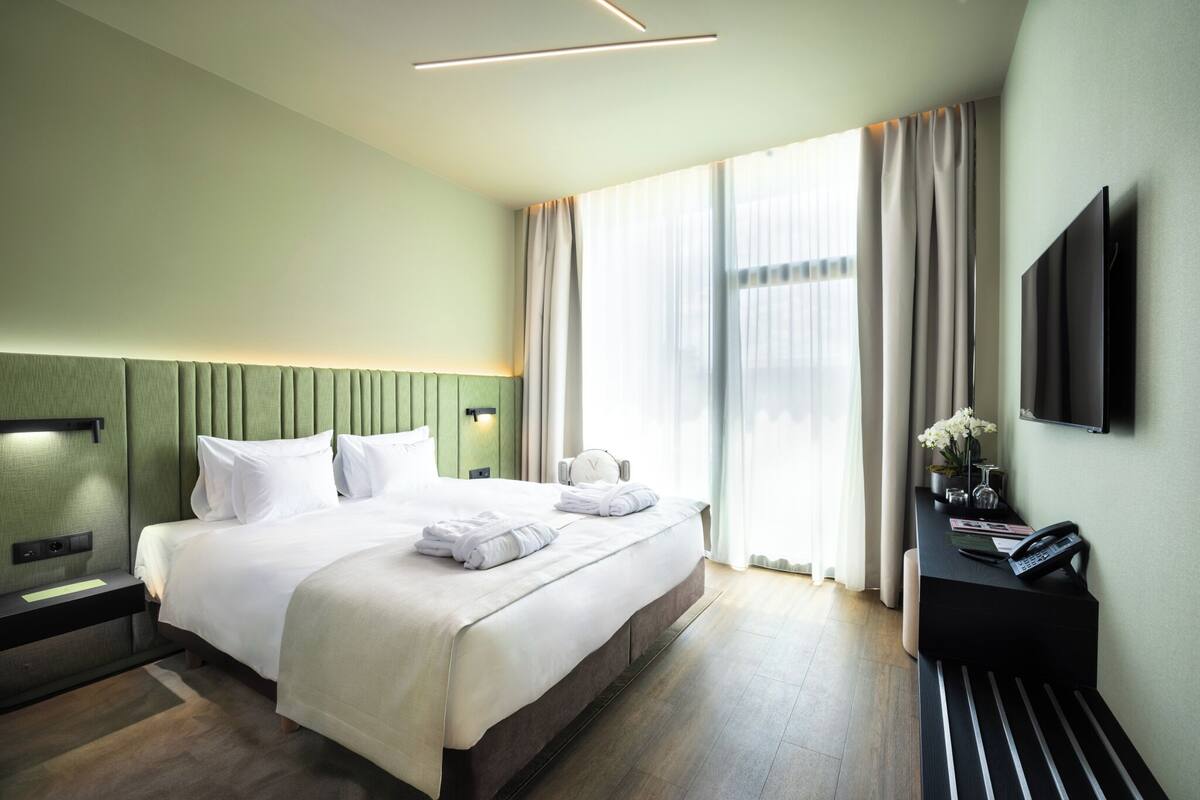 Una habitación de hotel moderna con una cama grande, mesitas de noche, un escritorio con silla, un televisor de pantalla plana y un amplio ventanal con cortinas.