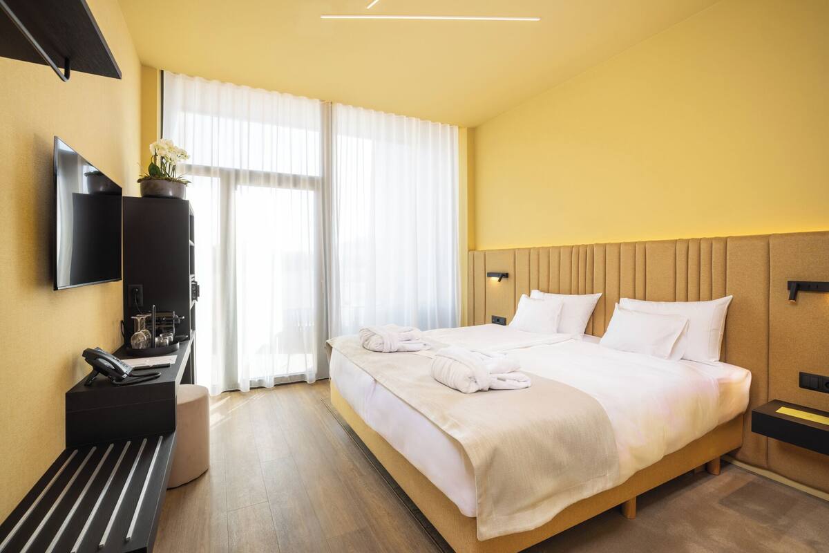 Habitación de hotel con una cama, un escritorio con teléfono, un televisor montado en la pared y una ventana con cortinas.