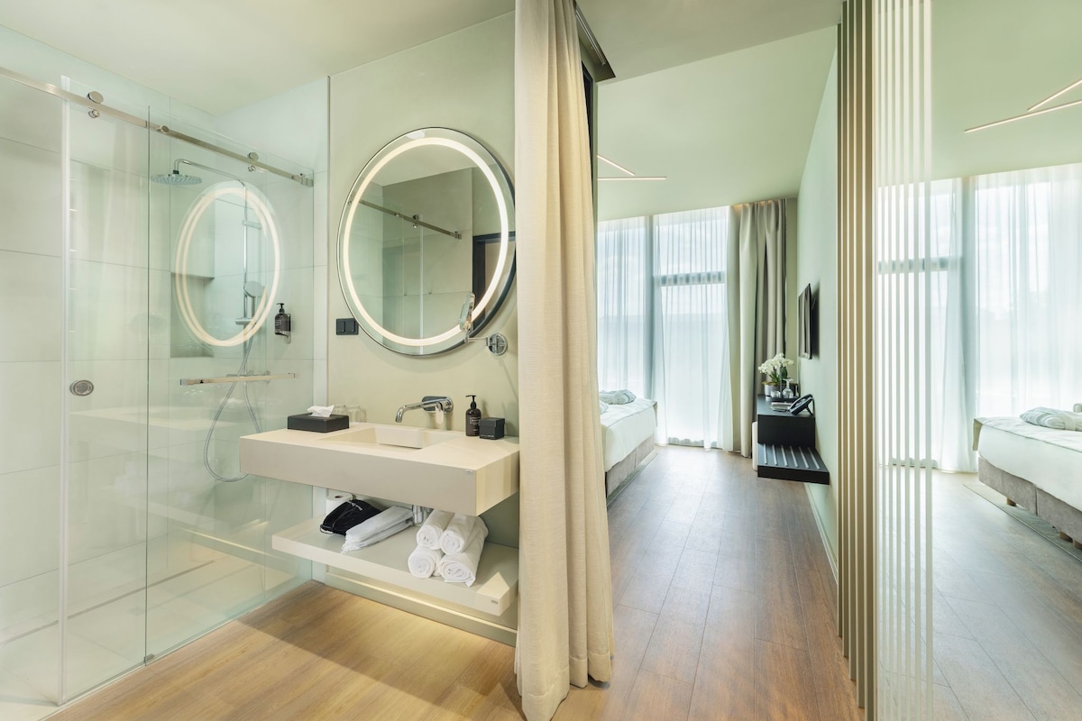 Une salle de bain moderne avec une douche à parois de verre, un meuble-lavabo avec un miroir et un lit dans la pièce adjacente.