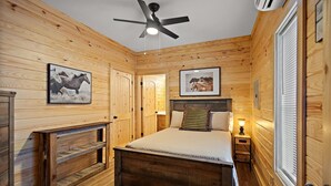 Camera da letto con rivestimento in legno, un letto, una ventola a soffitto e due quadri raffiguranti cavalli.