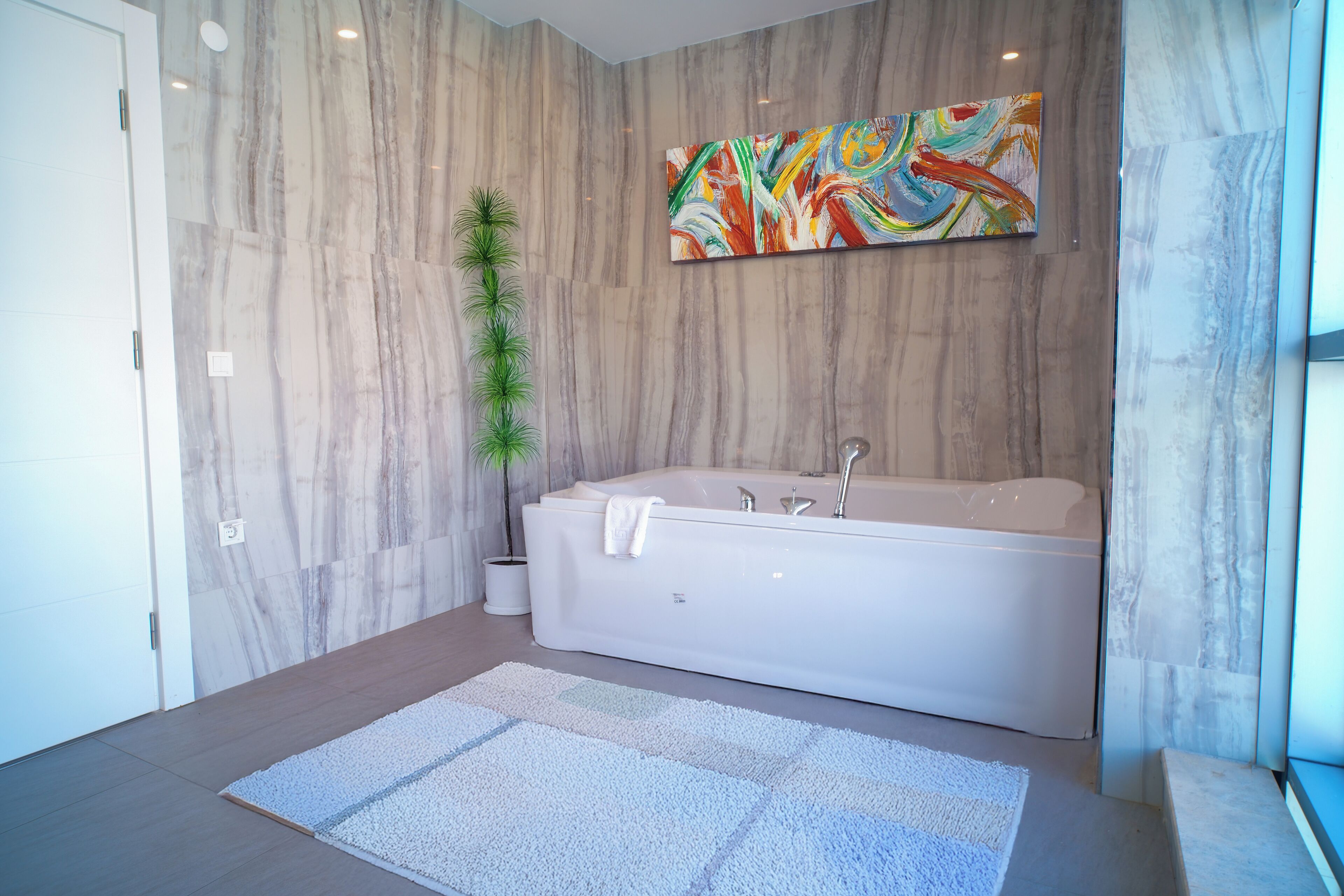 Un baño moderno con una bañera grande, paredes de mármol, un cuadro abstracto de colores y una planta en maceta.