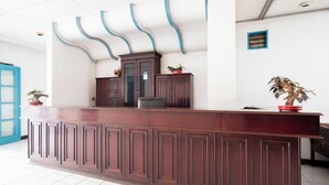A reception desk with a dark wood finish, a blue-framed window, and a potted plant on the counter. - Sartika Hotel (Bandung)