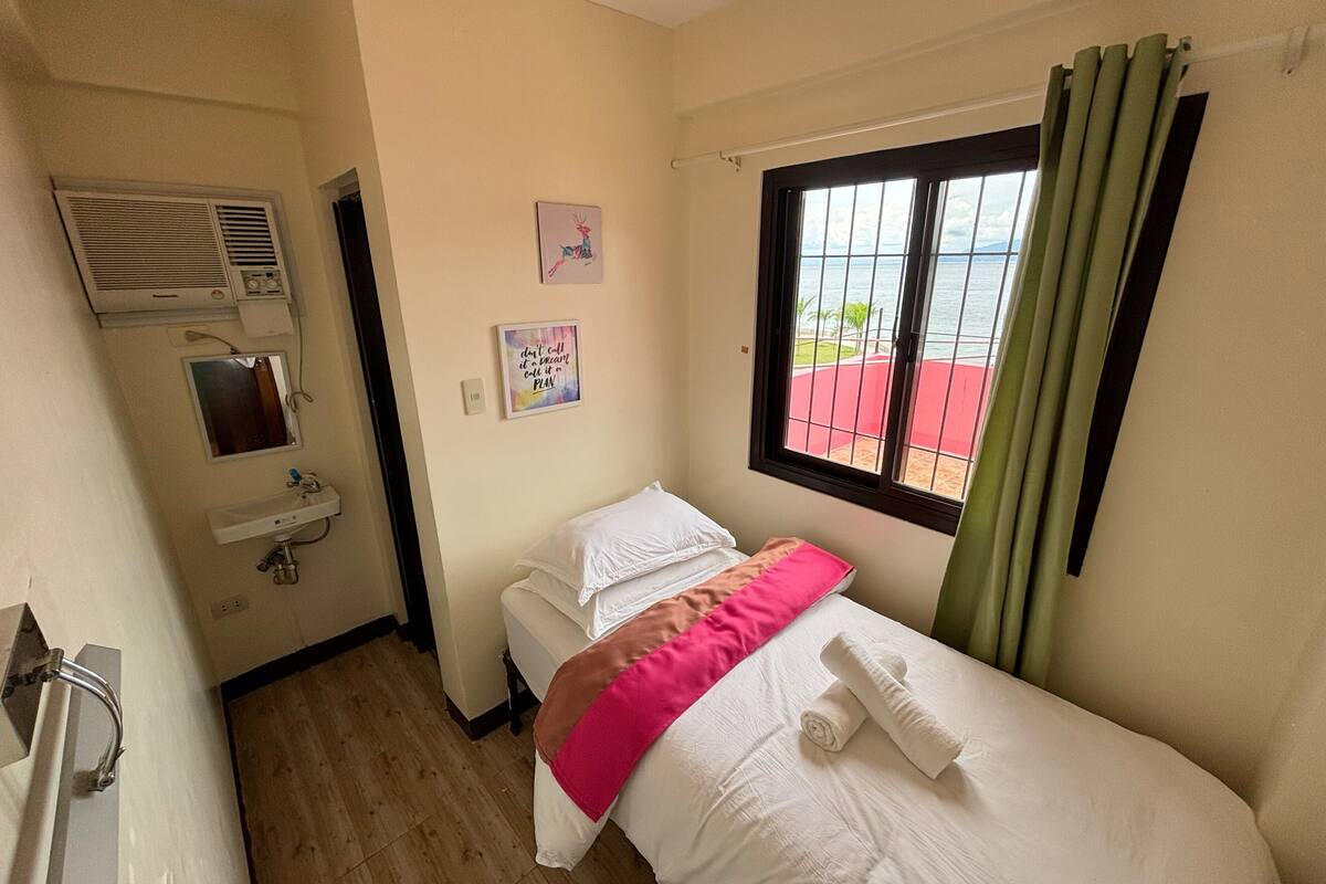 Una habitación pequeña y limpia con cama, ventana con cortinas, lavamanos y aire acondicionado instalado en la pared.