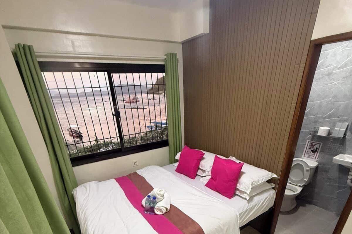 Habitación de hotel con cama, ventana con cortinas, vista a la playa y baño con inodoro y lavabo.