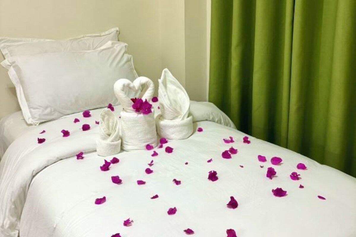 A neatly made bed with white linens and a heart-shaped towel arrangement, green curtains, and scattered rose petals.