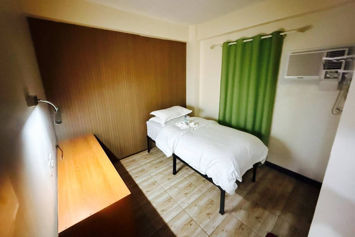 Una habitación pequeña con cama individual, piso de madera, lámpara de noche, cortina verde y equipo de aire acondicionado.