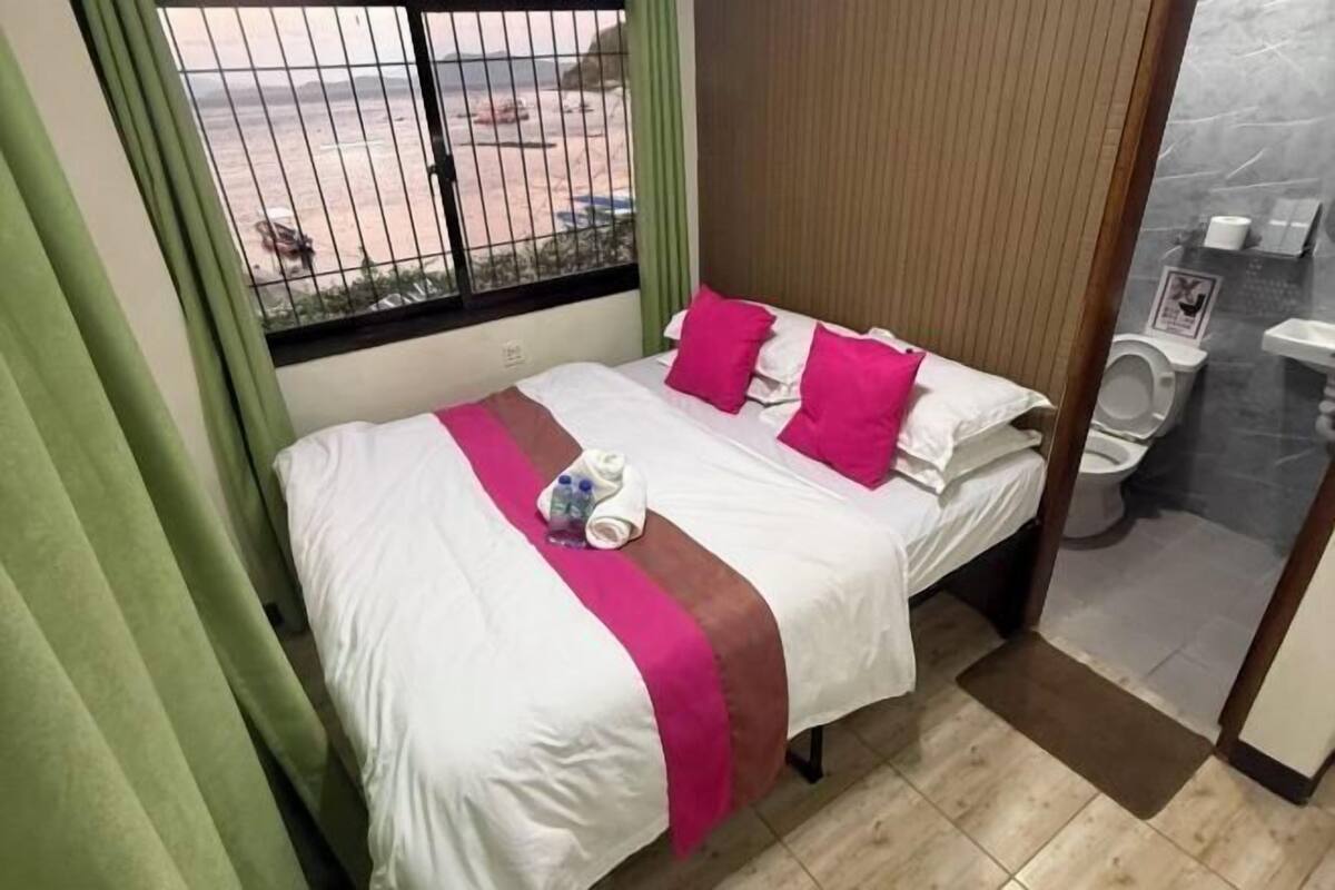 Habitación de hotel con cama, ventana con vistas, baño con inodoro y lavamanos, y cortina verde.