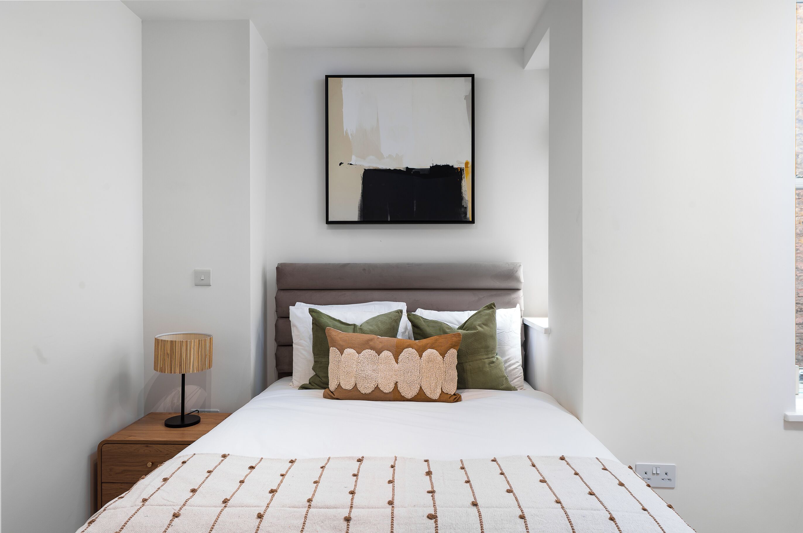 Une chambre à coucher avec un lit, une table de chevet, une lampe et une œuvre d’art encadrée accrochée au mur.