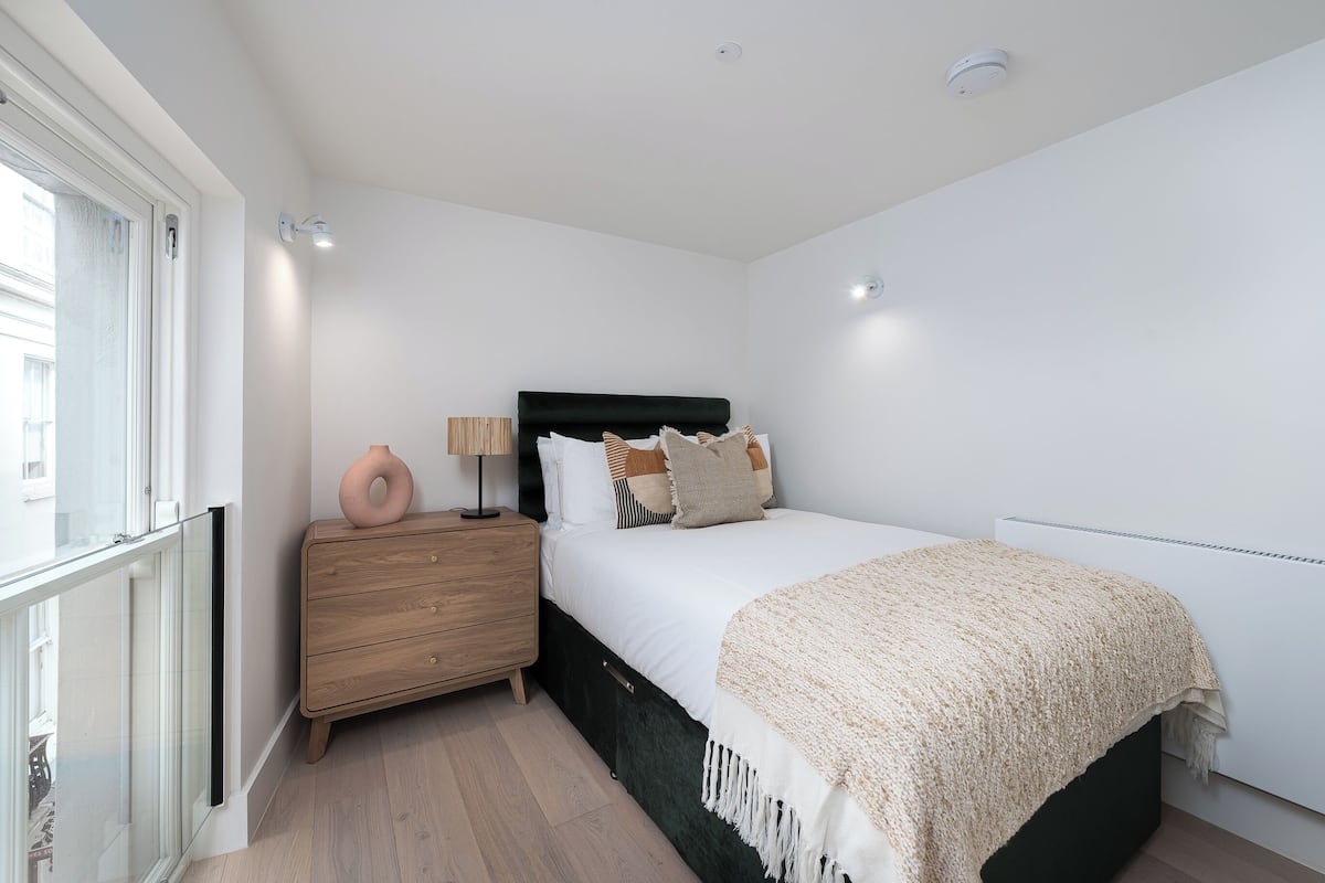 A bedroom with a bed, bedside table, lamp, and a view of the outside.