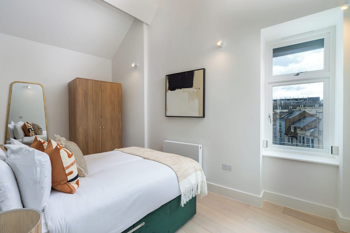 A bedroom with a bed, a wardrobe, a radiator, a window with a city view, and a framed artwork on the wall.