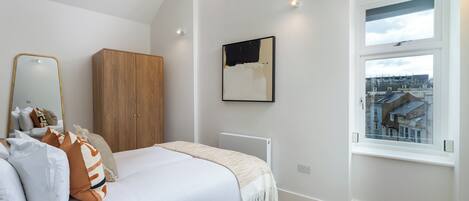 A bedroom with a bed, a wardrobe, a radiator, a window with a city view, and a framed artwork on the wall.