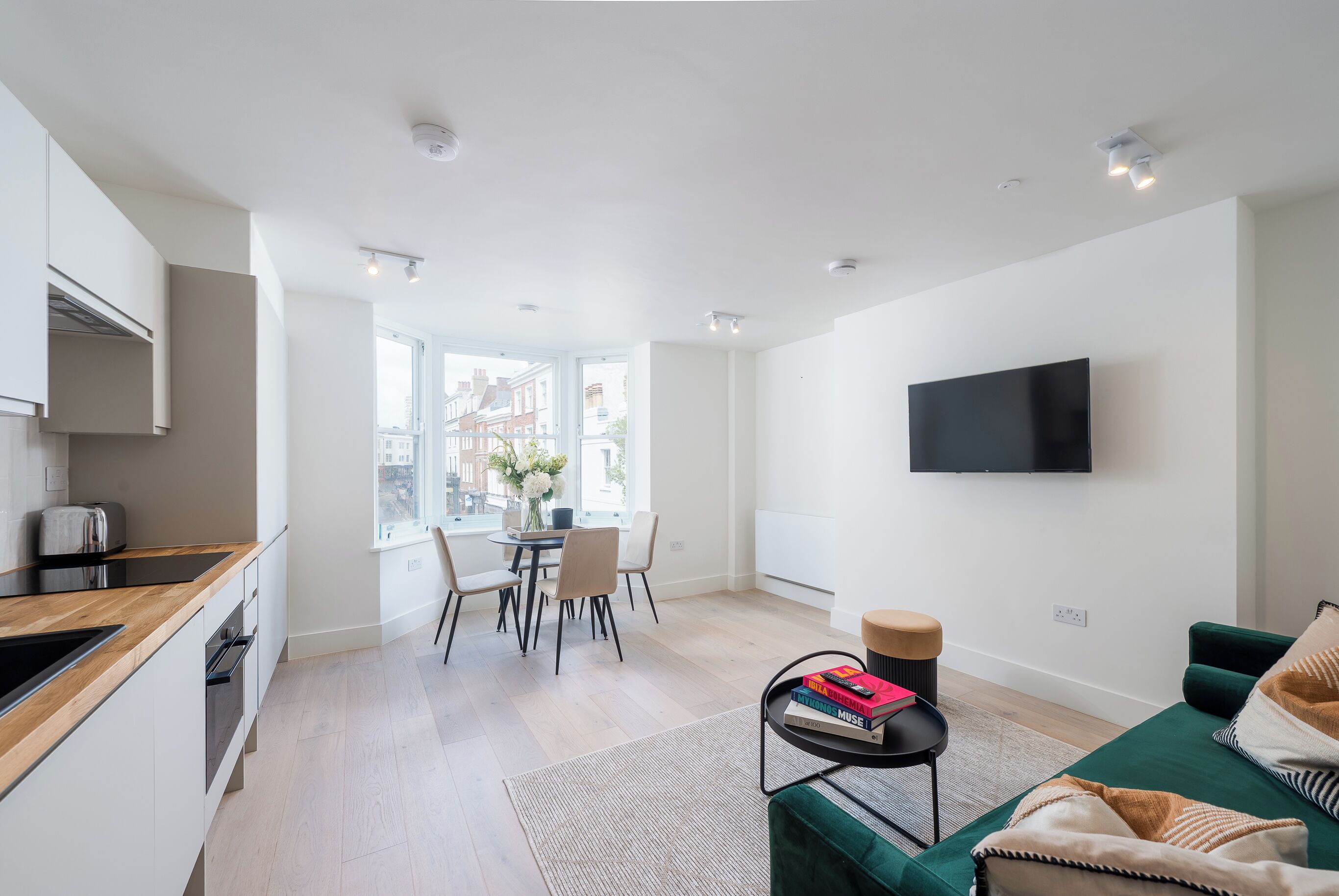 A modern apartment with a kitchen, dining area, and living room. It features a flat-screen TV mounted on the wall, a wooden kitchen counter, and a green sofa.