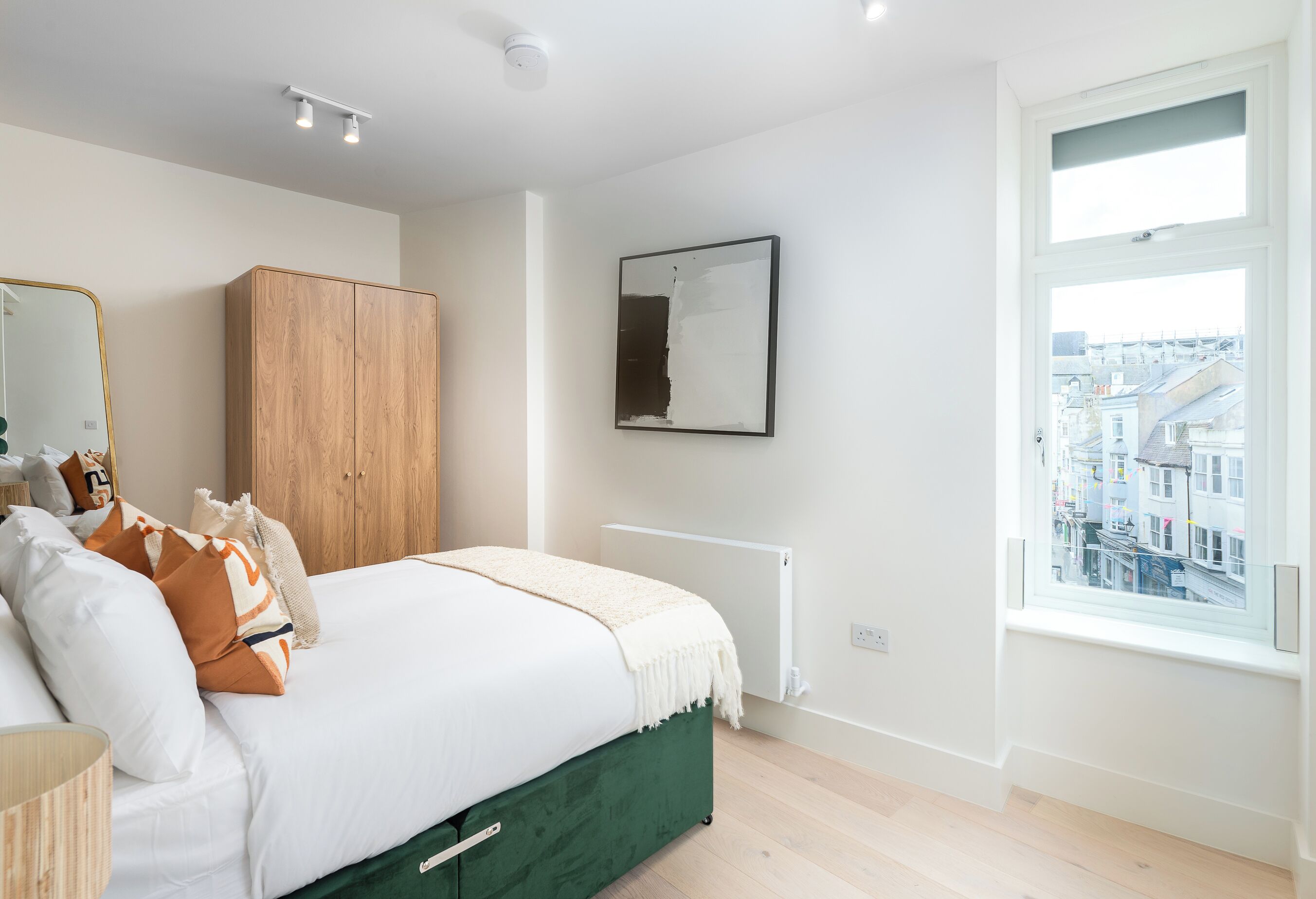 Un dormitorio con cama, armario de madera, espejo, radiador y ventana con vista a la ciudad.