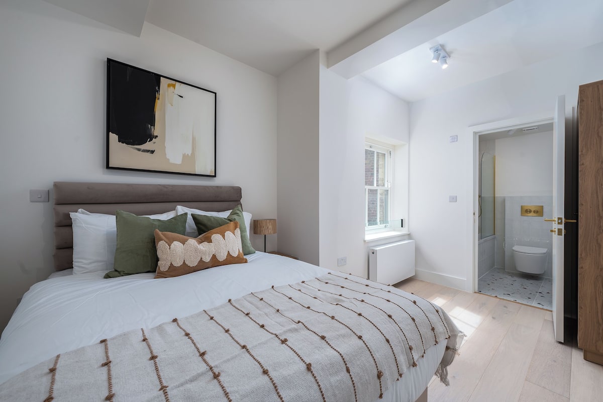 A bedroom with a large bed, a bedside table, a window, and a door leading to a bathroom.