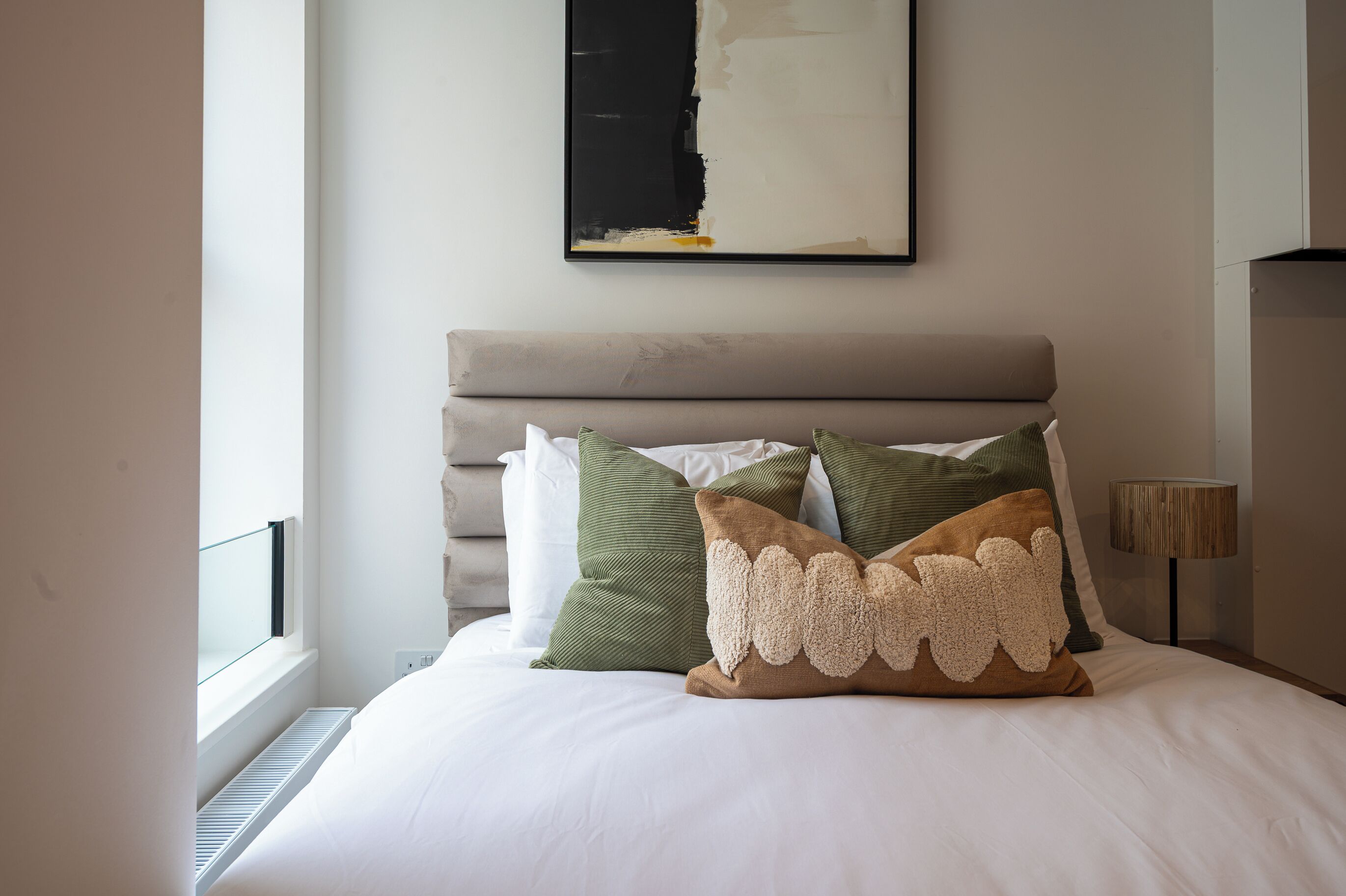 A neatly made bed with a headboard, a window with blinds, and a wall-mounted abstract painting.
