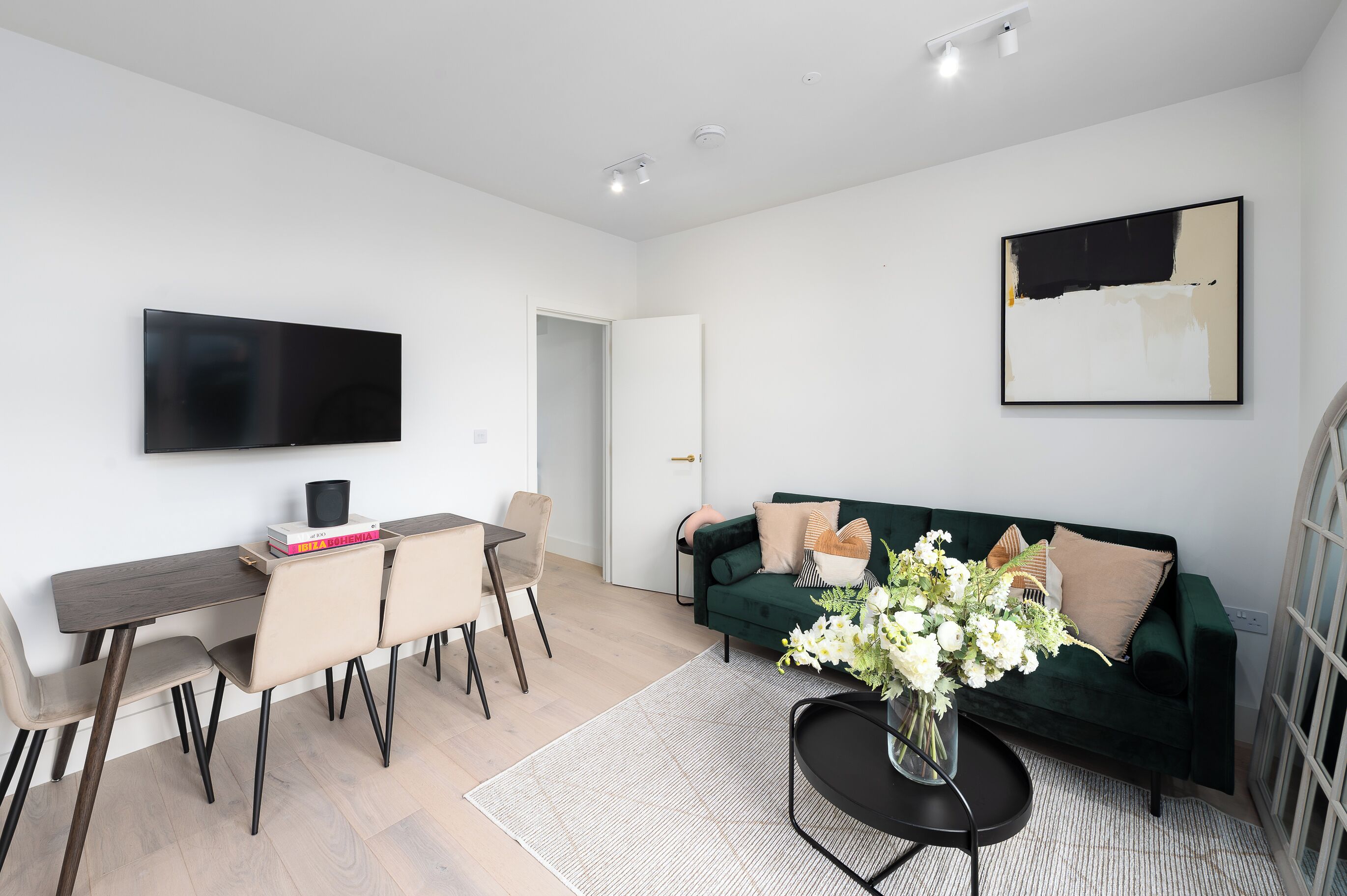 Un salon moderne avec un téléviseur à écran plat, un canapé vert foncé, une table à manger avec des chaises et une table basse ornée d’un vase de fleurs.