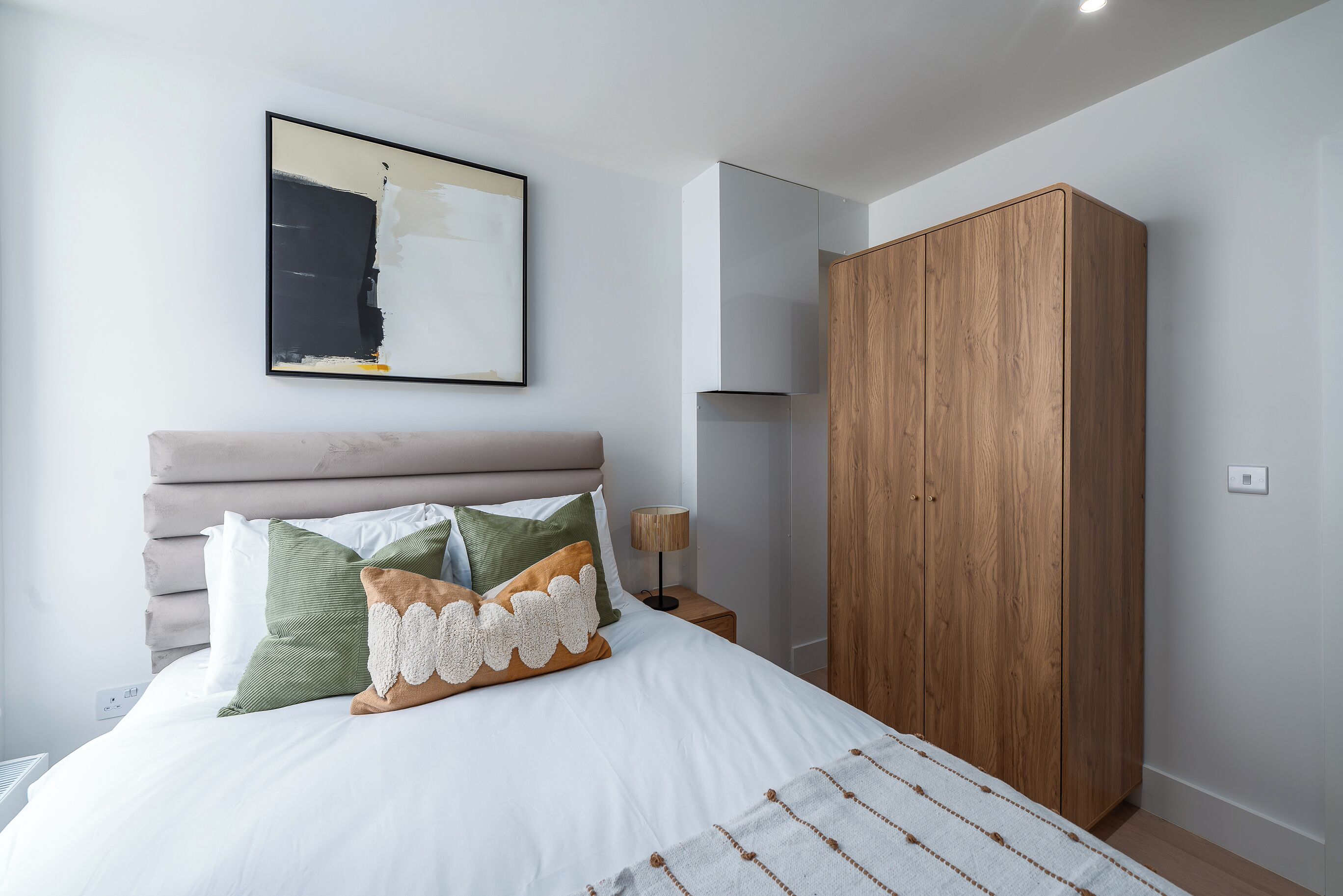 A bedroom with a large bed, a wooden wardrobe, a nightstand with a lamp, and a painting on the wall.