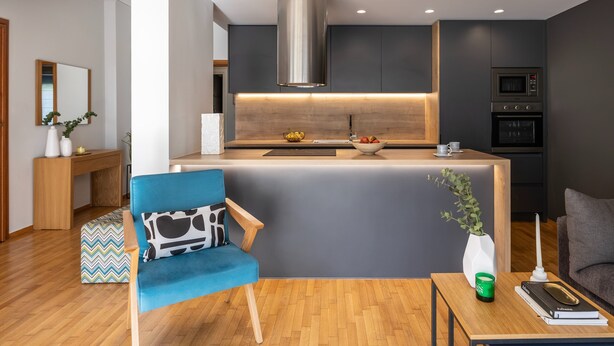 A modern kitchen with a central island, a built-in microwave, and an oven. There is a blue chair with a patterned cushion, a small table with a book and a green candle, and a potted plant.