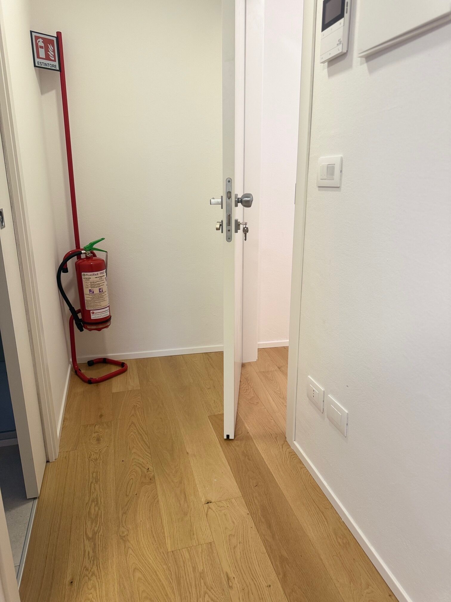 Un pasillo con suelo de madera, una puerta blanca, un extintor colgado en la pared y un termostato en la pared.