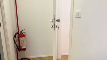 Un pasillo con suelo de madera, una puerta blanca, un extintor colgado en la pared y un termostato en la pared.