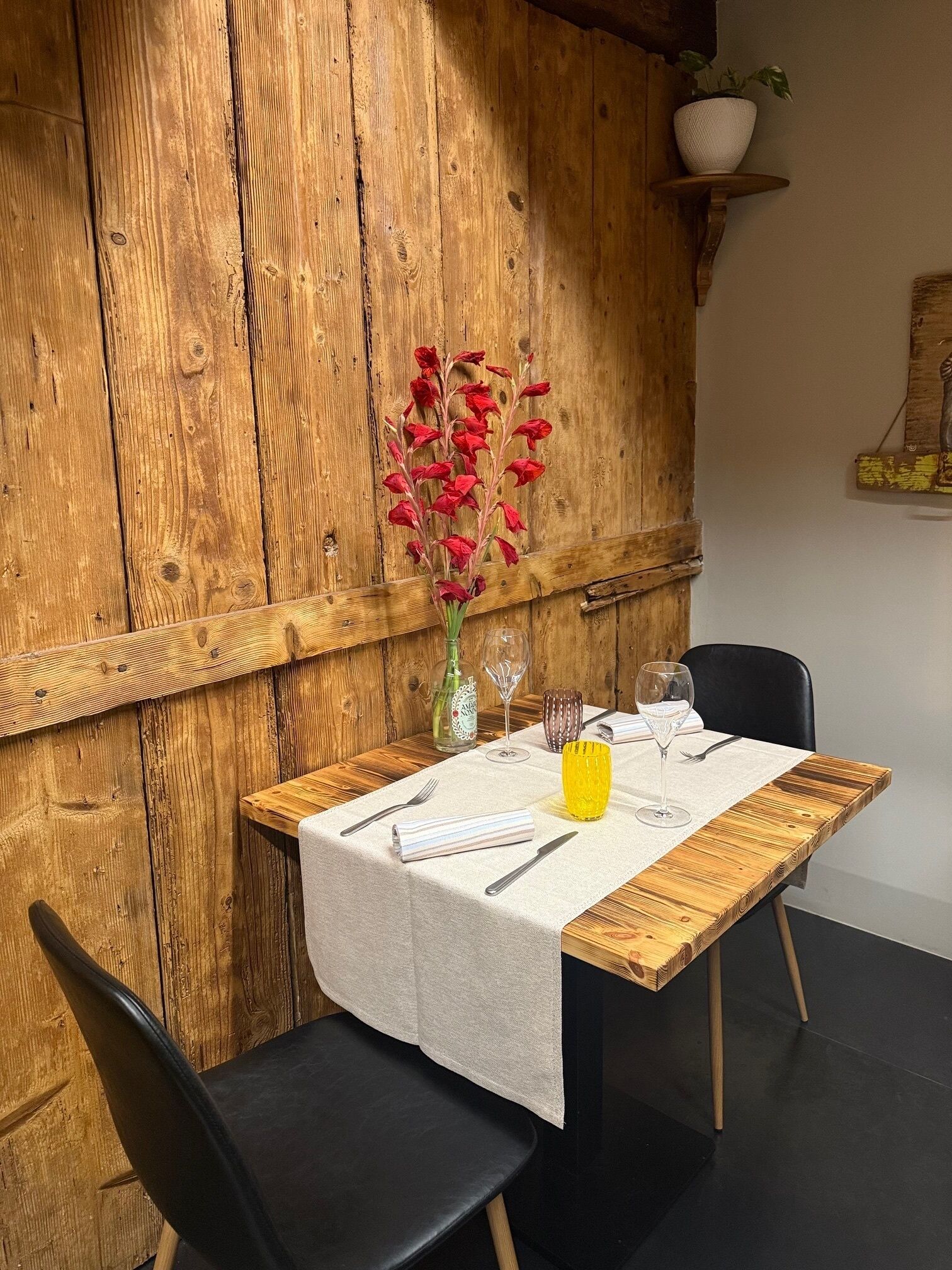 Uma mesa de jantar de madeira com toalha branca, duas cadeiras pretas e um vaso com flores vermelhas.
