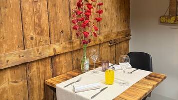 Uma mesa de jantar de madeira com toalha branca, duas cadeiras pretas e um vaso com flores vermelhas.