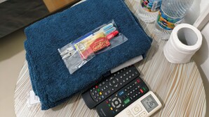 A neatly made bed with a striped pillow, a blue towel, two water bottles, a remote control, and a roll of toilet paper on a small round table. - Ez Door (Yogyakarta)