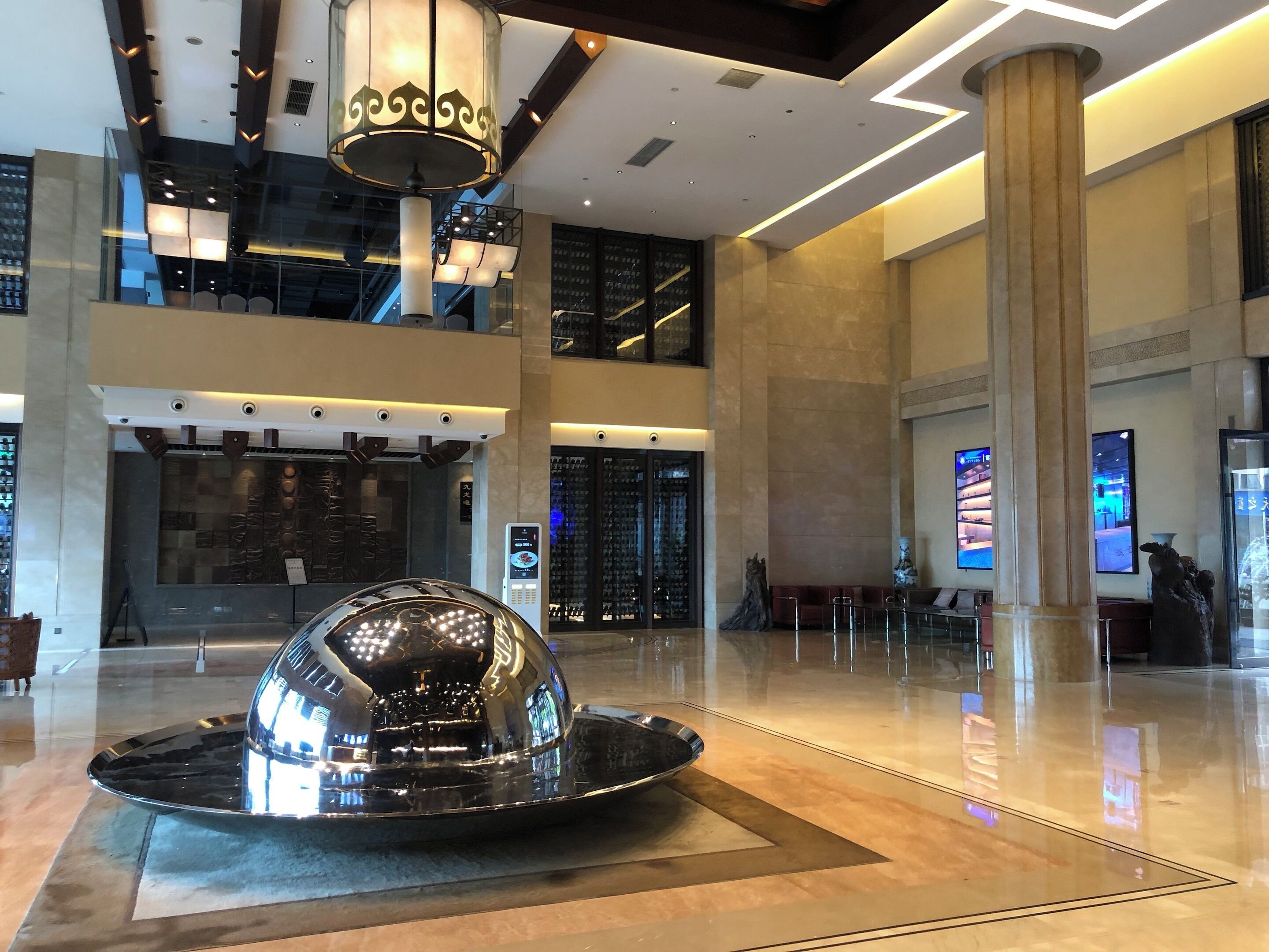 A spacious lobby with a central reflective sculpture, modern lighting fixtures, and a digital display.