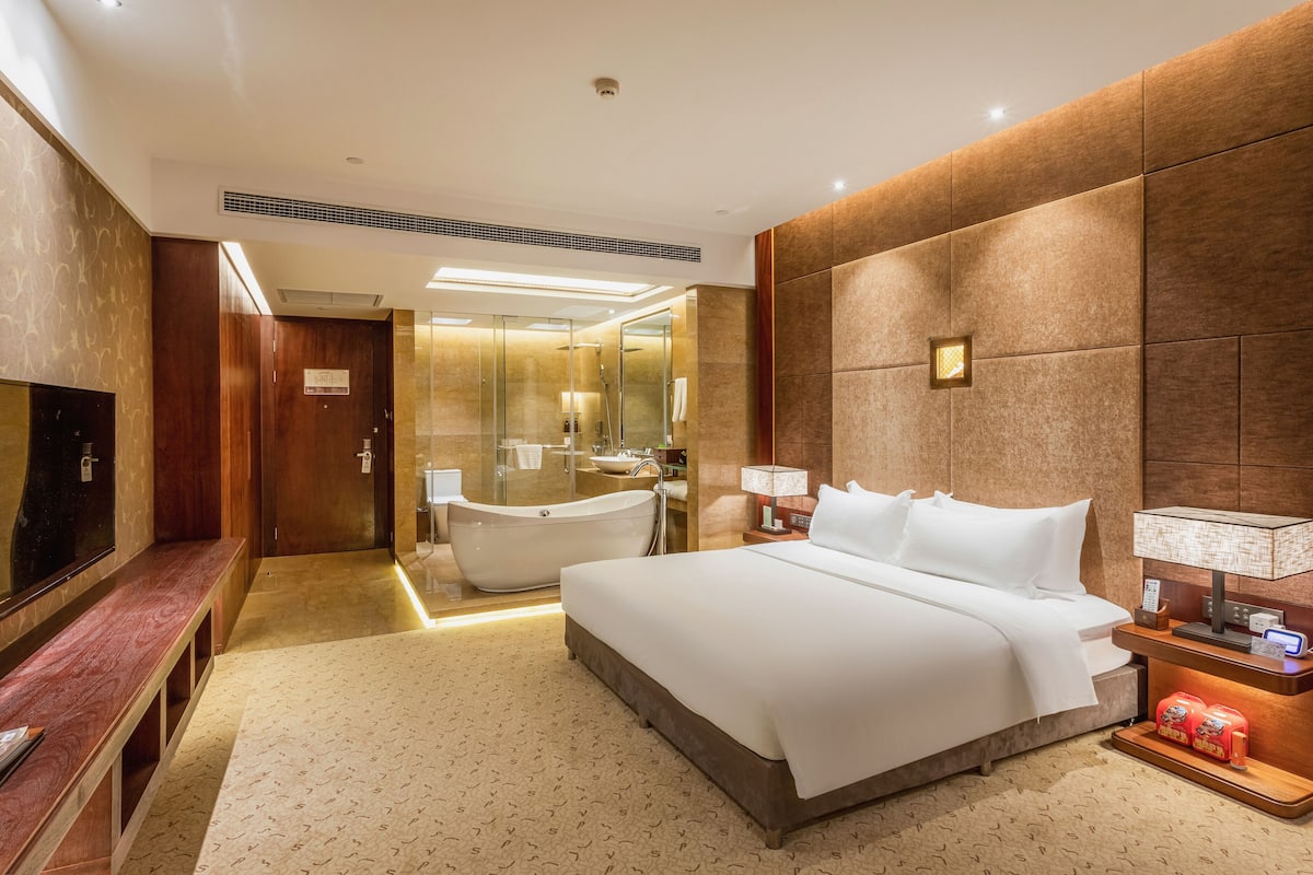 A modern hotel room with a large bed, a television, a bathtub, and a bathroom with a glass shower enclosure.
