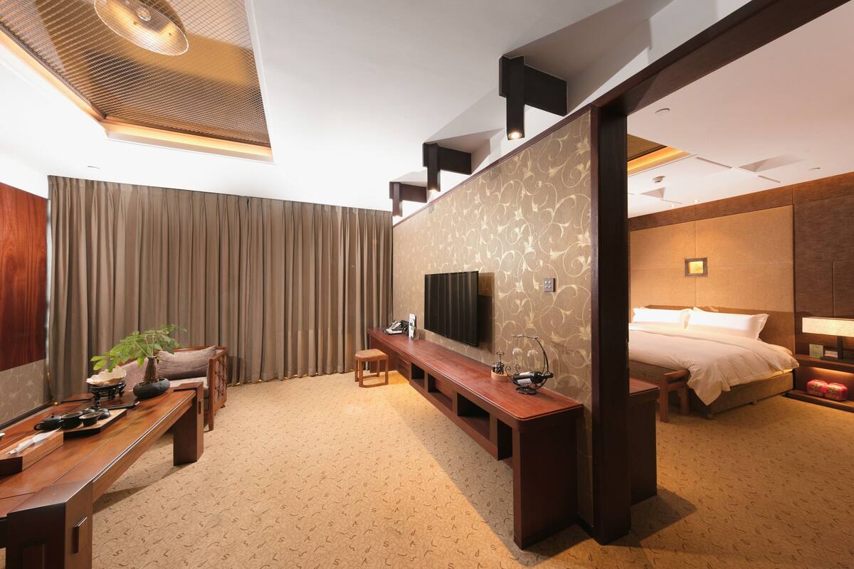 A modern hotel room with a large bed, a TV, a wooden bench, and a desk with a plant.