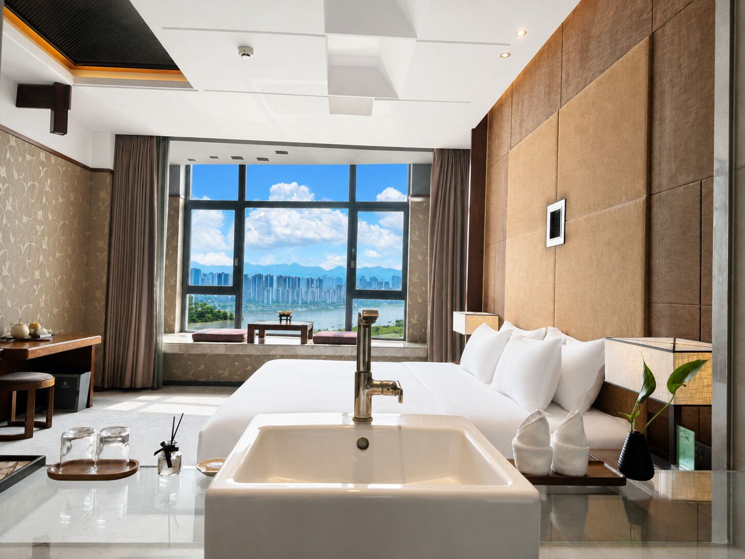 Habitación de hotel moderna con una cama grande, un baño con lavamanos blanco y vistas a un paisaje urbano a través de amplios ventanales.