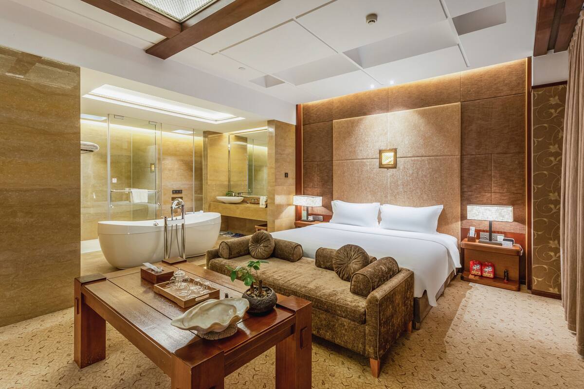 A modern hotel room with a large bed, a wooden table with amenities, a bathtub, and a well-lit bathroom.
