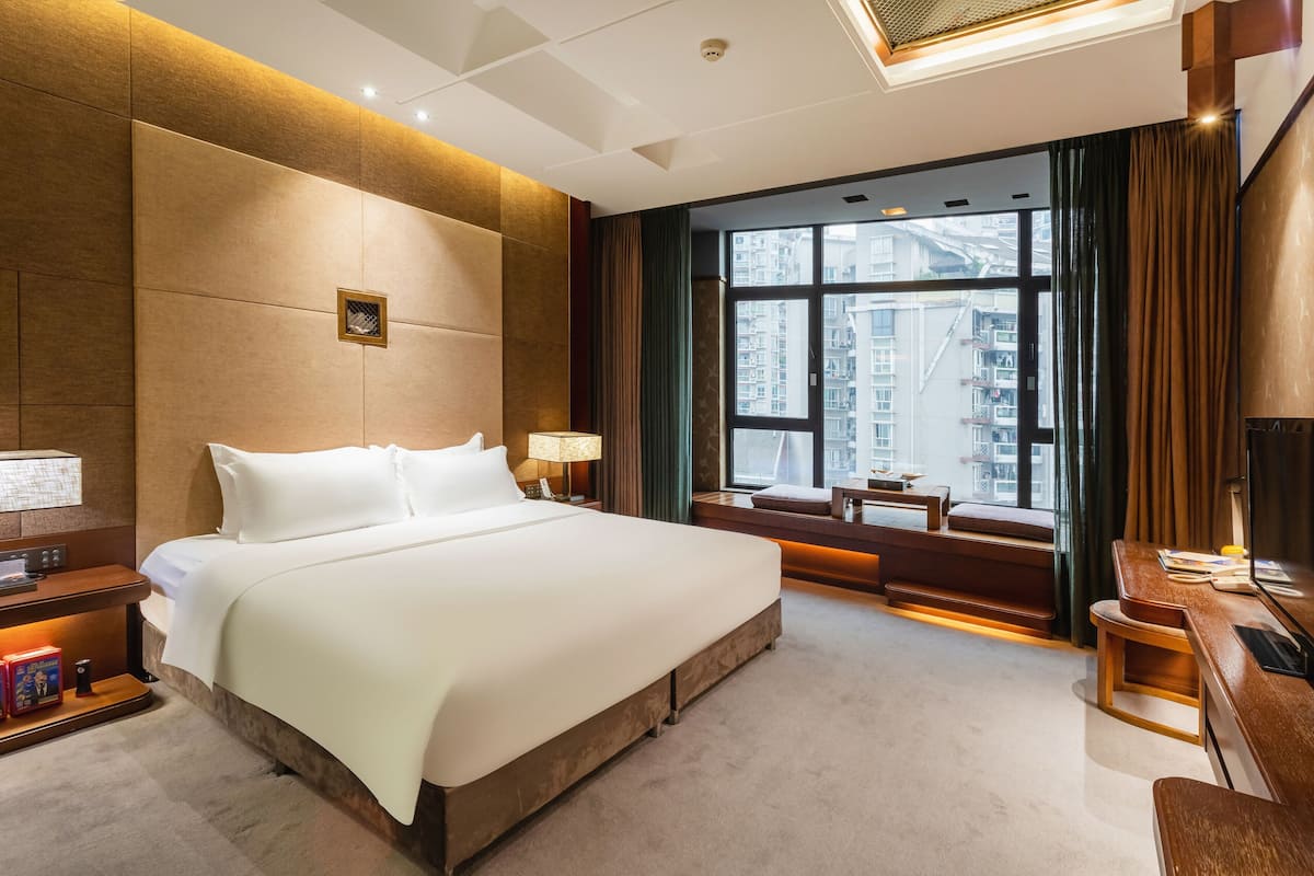 A modern hotel room with a large bed, a desk, and a view of the city through large windows.