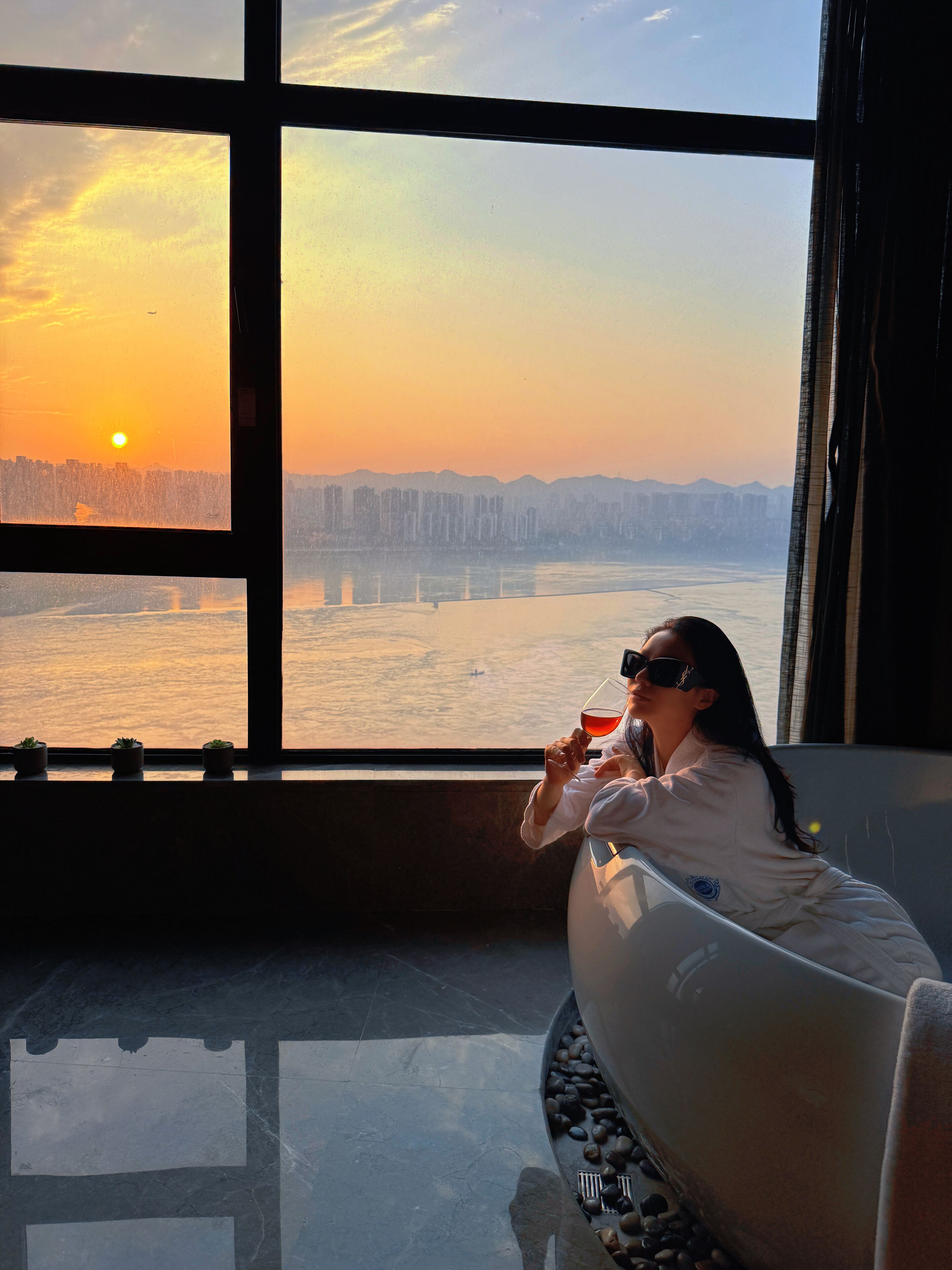 A person sitting by a window with a view of a city skyline and a body of water during sunset.